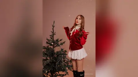 Cheerful woman in a red velvet top and white skirt standing by a decorated Christmas tree in a festive setting.