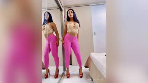 Young woman in a beige lace crop top and bright pink leggings posing in a bright bedroom mirror.