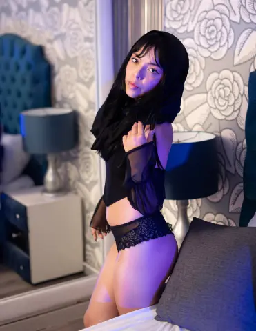 Young woman in black lace lingerie and sheer top posing in a bedroom with floral wallpaper.