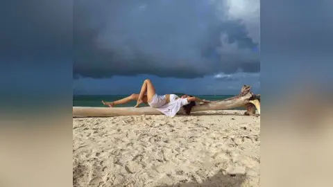 Relaxed woman in white loungewear lying on driftwood on a sandy beach under a moody cloudy sky.
