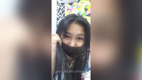 Smiling young woman with long black hair playing with her hair in a colorful indoor setting.