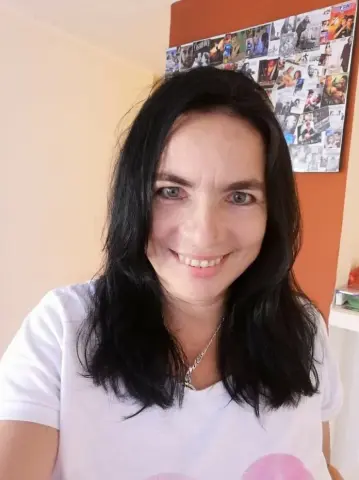 Smiling woman with long black hair wearing a white t-shirt in a bright indoor setting.