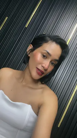 Smiling woman in a white strapless top posing against a dark textured wall background.
