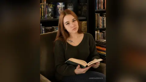 Redheaded woman in a dark green sweater reading a book in a cozy home library setting.