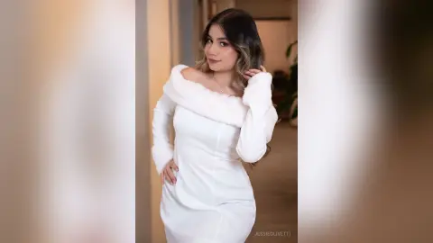JessieOlivetti poses in a white long-sleeved dress with a white fur collar, running her hand through her long brown hair.