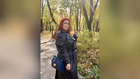 Redheaded woman in a black leather trench coat carrying a colorful crochet bag in a sunny autumn park.