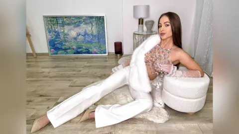 Glamorous woman in white flared trousers and a faux fur shrug sitting on a white pouf in a bright room.