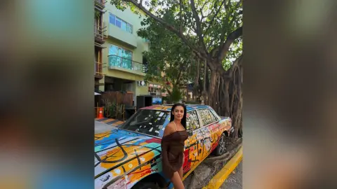 Stylish woman in a brown dress leaning on a colorful vintage car in a sunny urban street setting.