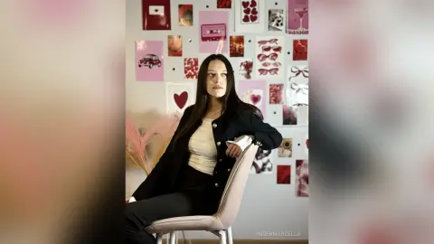 IngerMarzella poses sitting in a chair against a wall decorated with pink and red heart-themed posters, wearing a black cardigan over a white top.