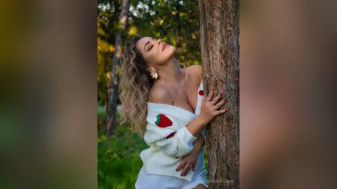 Blonde woman in a white off-the-shoulder sweater leaning against a tree in a lush green forest.