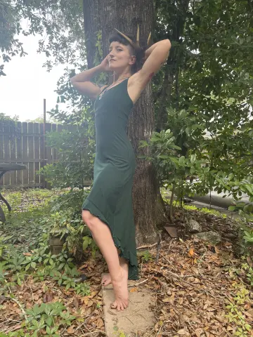 Young woman in a dark green maxi dress posing by a tree in a lush garden setting.