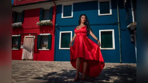 MissyJolie stands against a blue wall wearing a red satin high-low dress with a deep neckline.