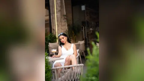 NicoleBella sits at a white table wearing a white sleeveless dress and sunglasses, looking down while holding a cocktail glass.