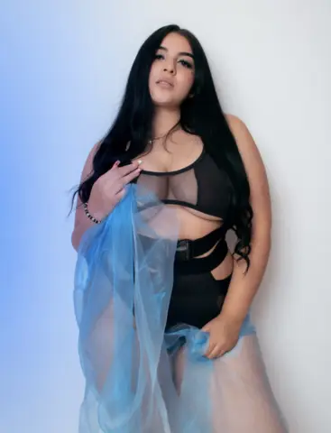 Young woman in black lingerie and sheer blue fabric posing against a soft blue studio background.