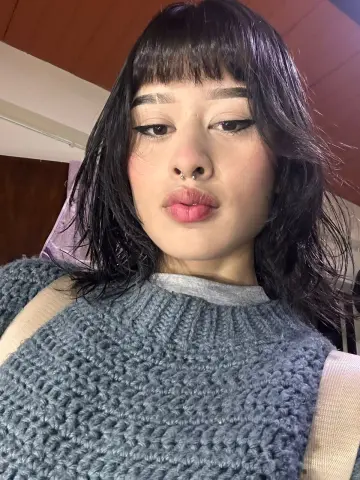 Young woman with dark bangs and nose piercing wearing a cozy grey knit sweater in a warm indoor setting.