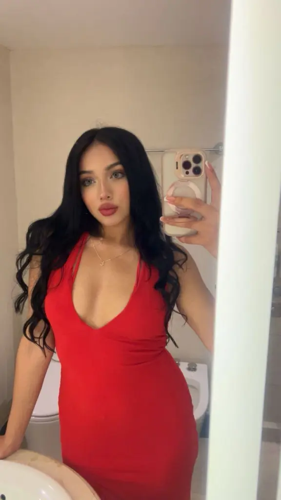 Young woman with long dark hair in a red low-cut dress taking a glamorous mirror selfie.