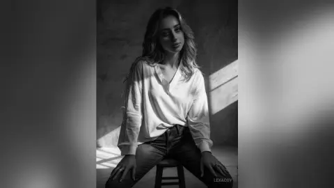 Moody black and white portrait of a blonde woman in a white long-sleeve shirt sitting in a sunlit room.