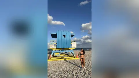 Young woman in a black bikini standing on Miami Beach sand next to a blue lifeguard tower under a sunny sky.