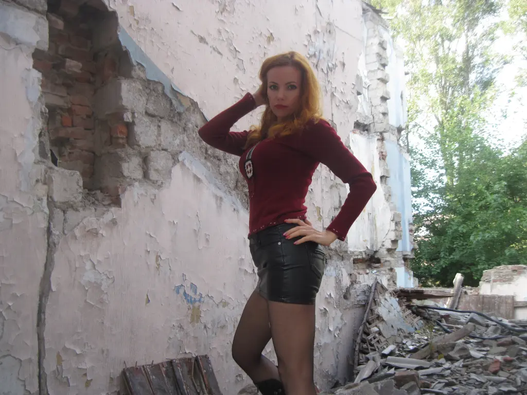 ElegantAndPretty poses with one hand on her hip, wearing a long-sleeved red top and black shorts against a weathered white wall.