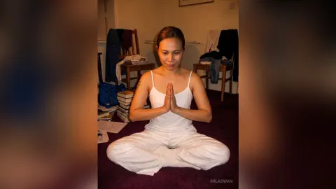 MilaSwan — MilaSwan sits cross-legged on the floor in a white tank top and white pants with her hands pressed together in prayer.