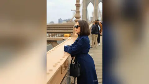 Stylish woman in a navy blue trench coat leaning on a bridge railing against a city skyline backdrop.