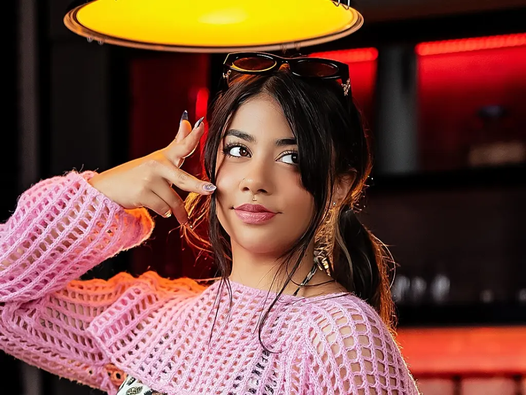 AnyaFord poses with a hand near her face, wearing a pink mesh top with her dark hair styled down.
