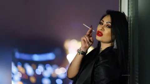 DeeaAnderson poses with a cigarette in her hand, wearing red lipstick and a black top against a dark, blurred background.