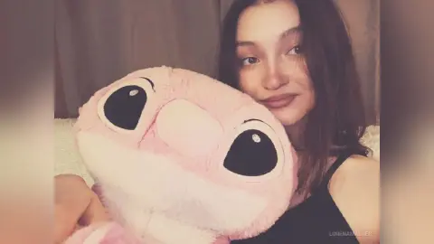Smiling young woman in a black tank top hugging a large pink Stitch plush toy in a cozy indoor setting.