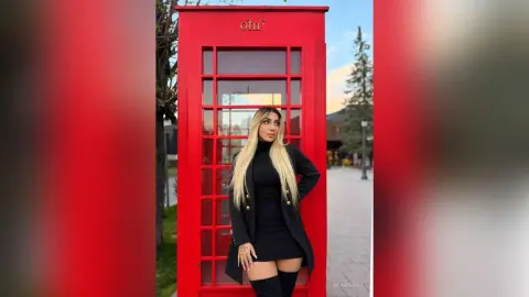Stylish blonde woman in a black dress and coat standing by a classic red British telephone booth outdoors.