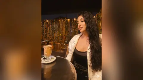 Smiling woman in a white jacket and black top enjoying coffee at a cozy outdoor cafe at night.