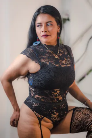 Confident woman in black lace bodysuit and large earrings posing in a bright indoor setting.