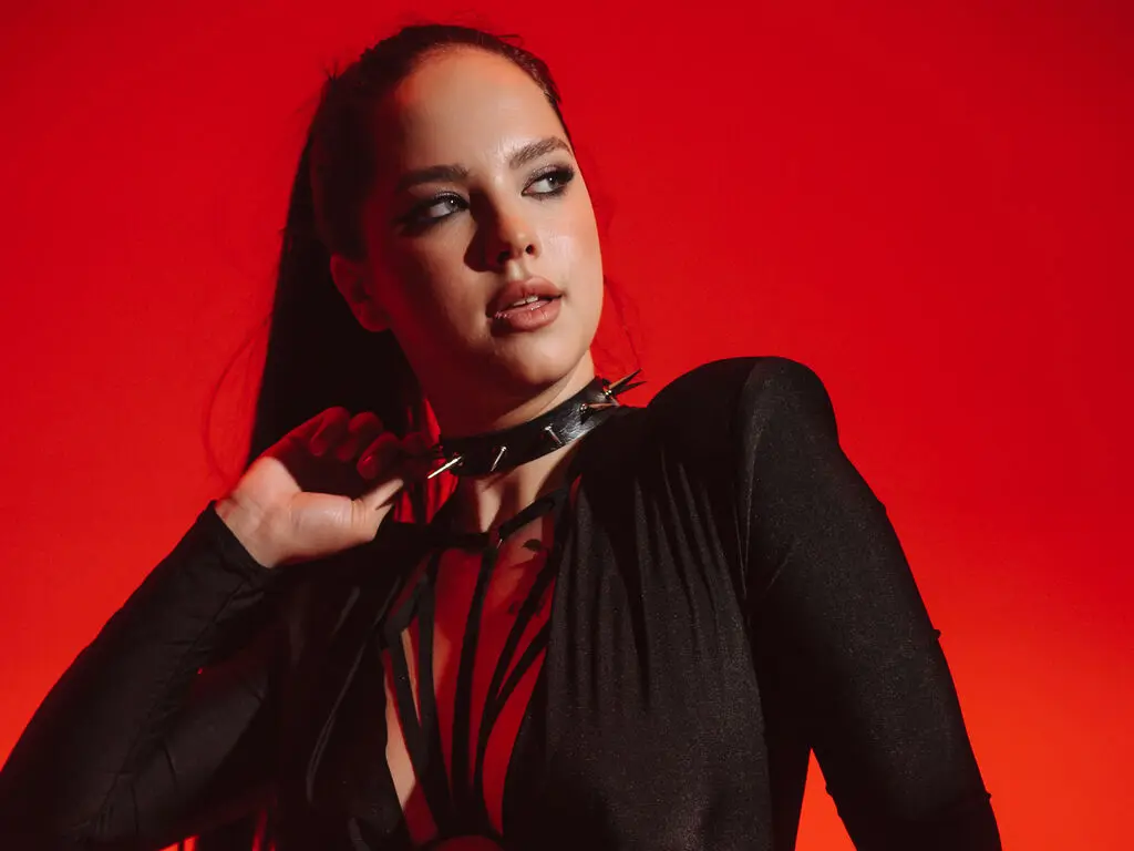 Edgy woman in a black long-sleeve top and spiked choker posing against a bold red background.