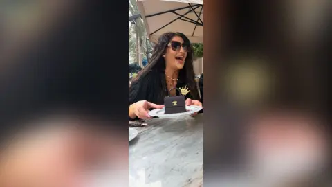 Excited woman in black outfit holding a small black Chanel handbag at an outdoor cafe table.
