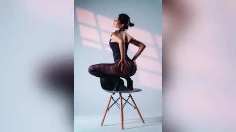 Elegant woman in a black bodysuit posing gracefully on a chair in a sunlit studio setting.