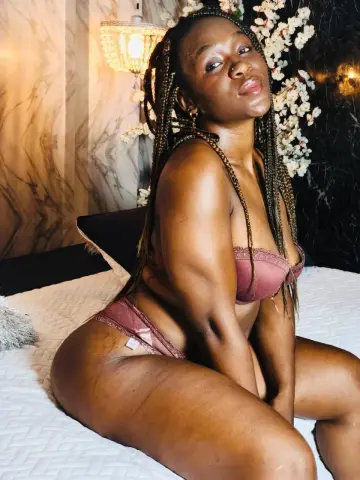 Confident Black woman with long braids in mauve lace lingerie relaxing on a white bed in a warm, moody setting.