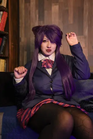Cosplayer in purple wig and school uniform sitting on a sofa with a moody expression.
