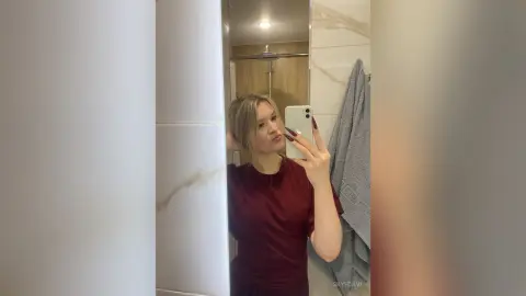 Young woman in a maroon t-shirt taking a casual mirror selfie in a modern bathroom.