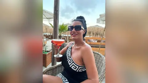 MissyJolie smiles while wearing black sunglasses and a black and white patterned tank top, holding a red cocktail in a glass.