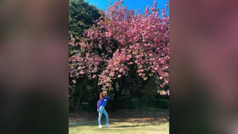 AnaMathers stands under a large, blooming pink cherry blossom tree in a sunny park wearing a blue top and light blue jeans.