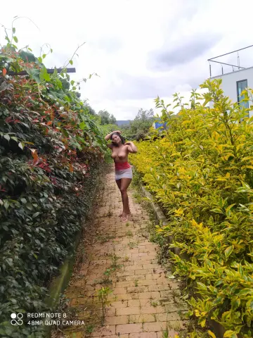 Young woman in a white crop top and red skirt standing on a brick path amidst lush green and yellow garden foliage.