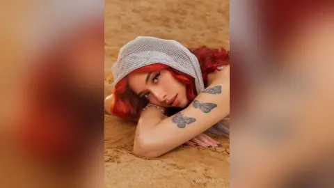 Red-haired woman with butterfly tattoos wearing a white mesh headscarf lying on a sandy beach in soft sunlight.