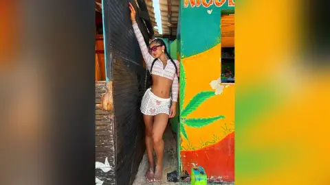 Stylish woman in a white crochet crop top and shorts leaning against a colorful wall in bright sunlight.