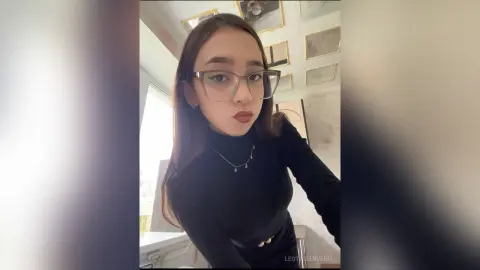 Young woman in a black turtleneck sweater and glasses posing in a bright, modern room.