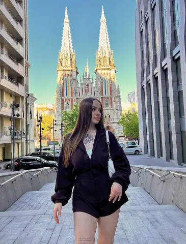 Young woman in a black romper standing on city stairs with a gothic cathedral in the background.