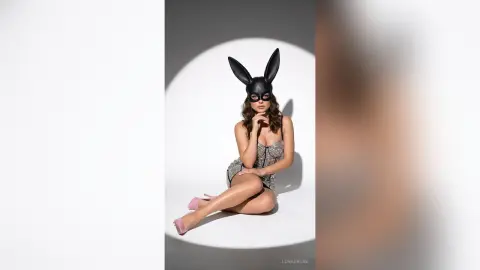 Glamorous woman in a sparkly silver bodysuit and black bunny mask sitting in a studio spotlight.