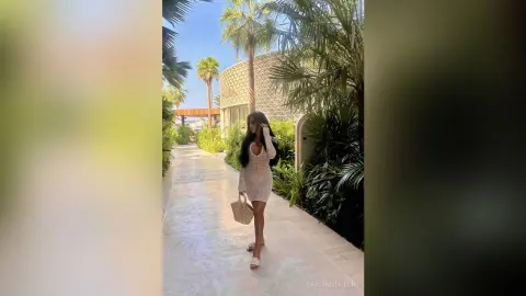 Stylish woman in a white crochet mini dress holding a straw bag walking through a tropical resort garden.