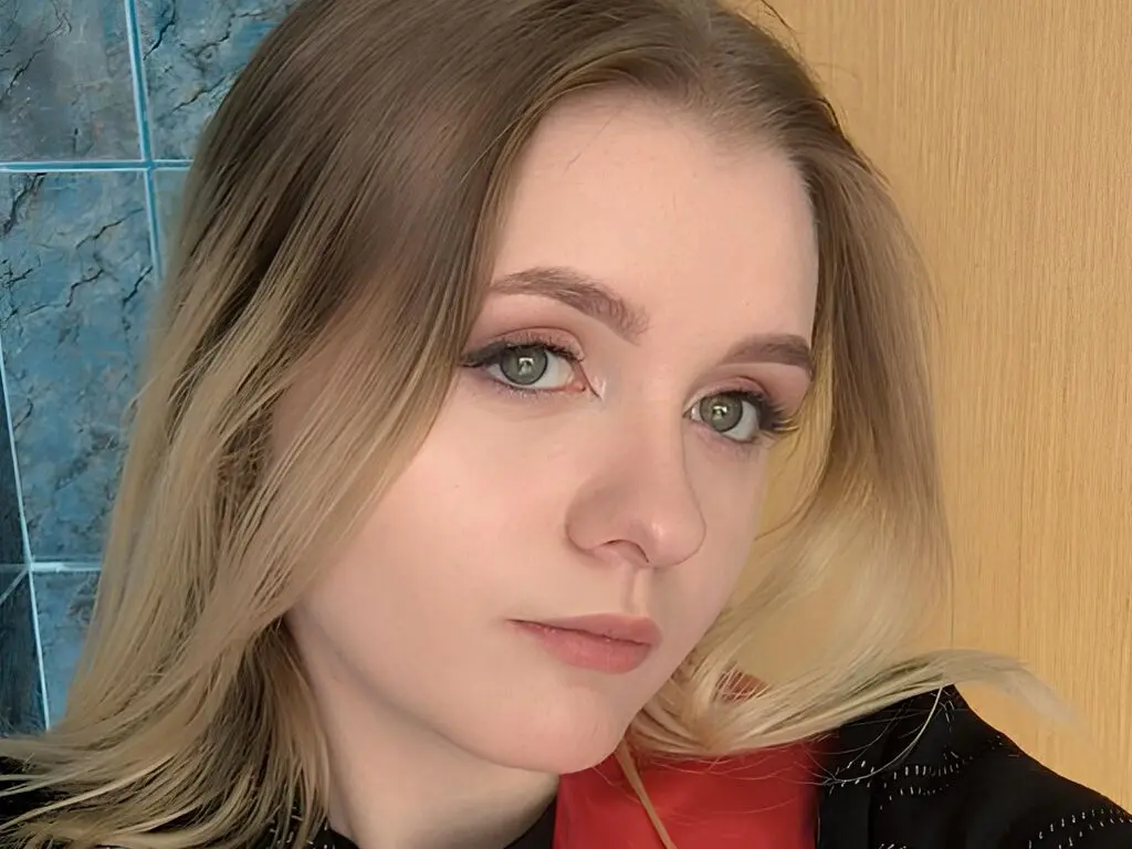 ShannonPomilla looks directly at the camera with blonde hair and light eye makeup against a blue tiled wall and wooden background.