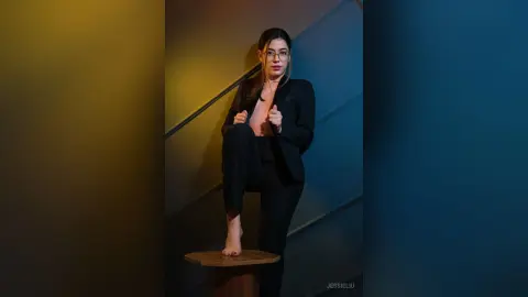 JessieLiu poses in a black suit and glasses, standing with one leg raised on a wooden stool.