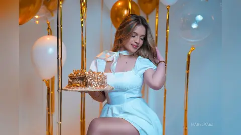 Smiling young woman in a white dress holding a gold cake at a celebratory birthday party with gold balloons.