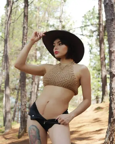 Young woman in a tan crochet halter top and black shorts posing in a sunny pine forest.
